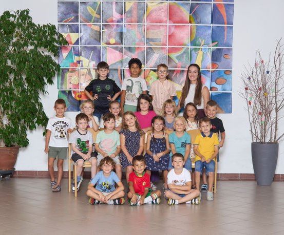Eine Gruppe von Kindern posiert für ein Foto in einem Klassenzimmer mit einer farbenfrohen Mosaikwand, Topfpflanzen und dekorativen Zweigen auf beiden Seiten.