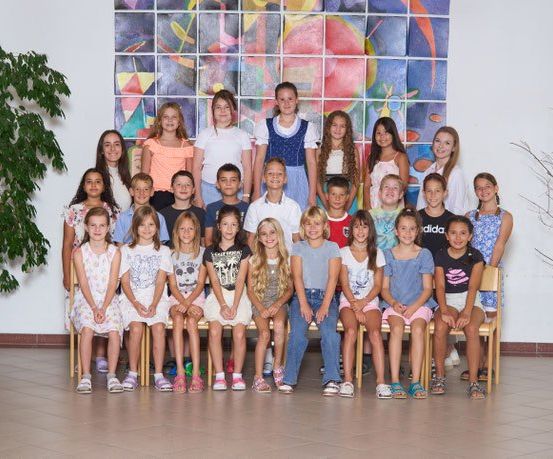 Ein Klassenzimmer voller Schüler, Jungen und Mädchen, posiert für ein Gruppenfoto vor einer Mosaikwand. Der Raum hat Topfpflanzen und einen gefliesten Boden.