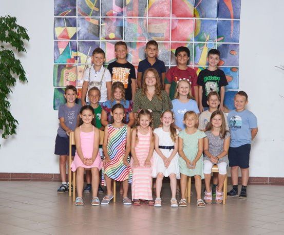 Eine Gruppe von Kindern und einer Frau posiert für ein Foto vor einer bunten Wand. Sie sitzen und stehen in zwei Reihen.