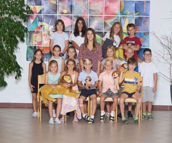 Eine Gruppe von Kindern und Erwachsenen, einschließlich einer Frau, posiert für ein Foto in einem Raum mit einer bunten Wand und Topfpflanzen.