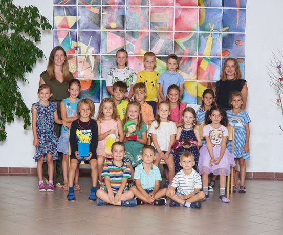 Eine Gruppe junger Kinder und zwei Frauen stehen vor einer bunten Wand und lächeln für ein Foto.