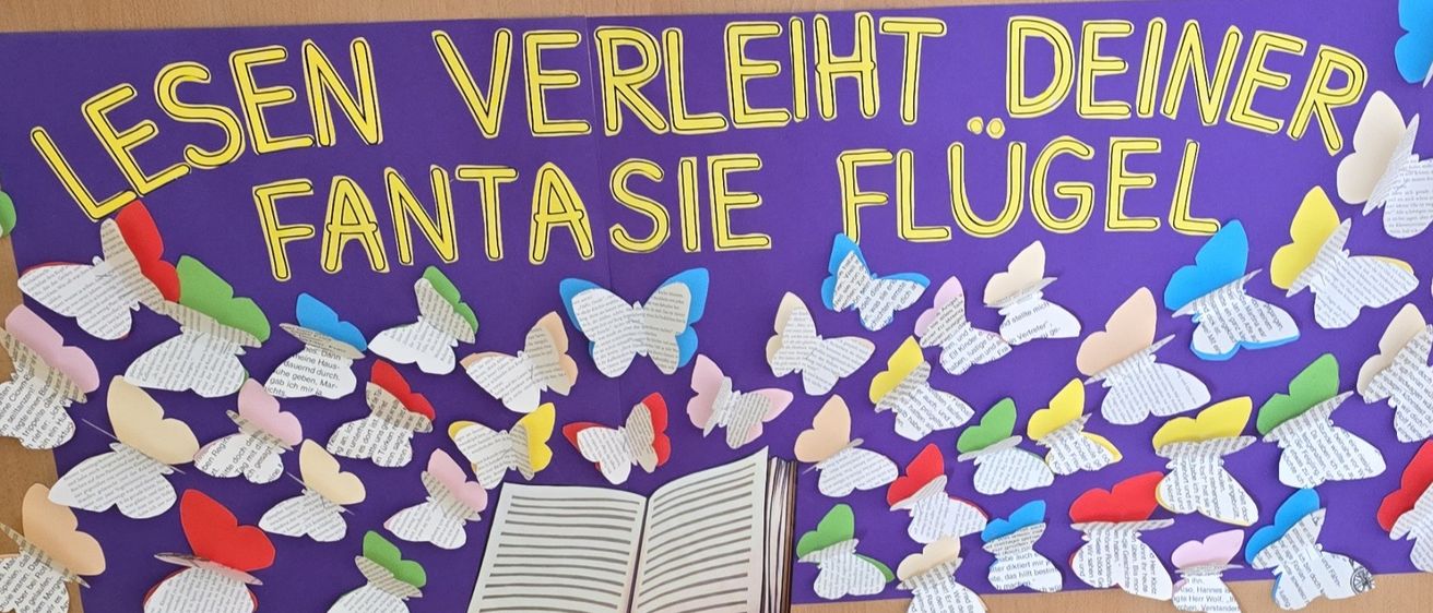 Ein farbenfrohes Bulletin Board zeigt einen deutschen Satz über Fantasie und Schmetterlinge, mit einem offenen Buch und zahlreichen farbigen Schmetterlingsausschnitten.