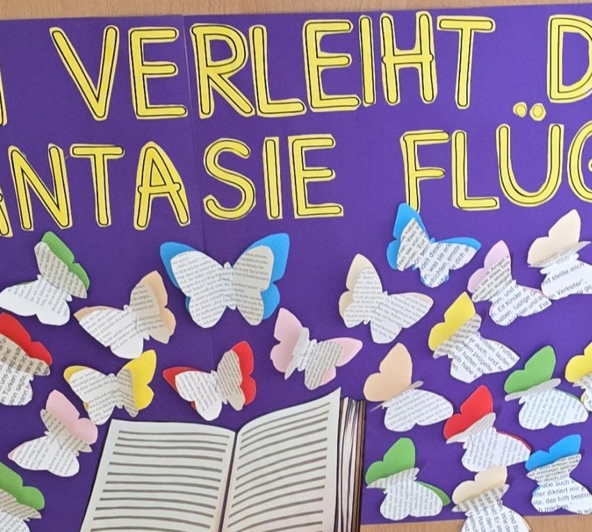 Ein farbenfrohes Bulletin Board zeigt einen deutschen Satz über Fantasie und Schmetterlinge, mit einem offenen Buch und zahlreichen farbigen Schmetterlingsausschnitten.