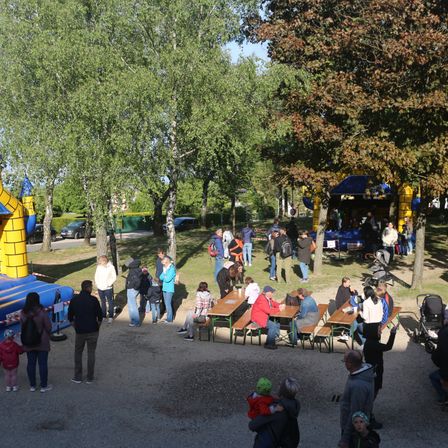 Ein Park mit Tischen, Bänken und einer gelben aufblasbaren Struktur. Menschen gehen herum, sitzen an Tischen und stehen in Gruppen. Es gibt Bäume und ein blaues Zelt.