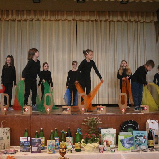 Mehrere Kinder in schwarzer Kleidung treten auf einer Bühne mit grünen Netzen auf, die vor einem weißen Vorhang stehen. Ein Tisch darunter hat Flaschen, Bücher und Blumen darauf.