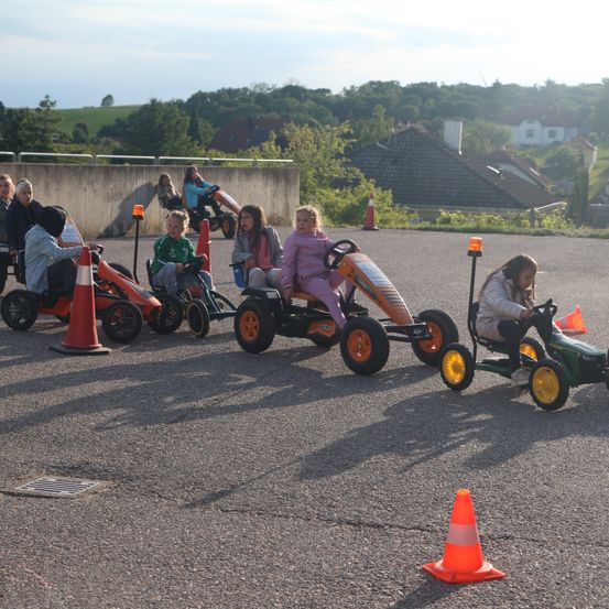 Kinder fahren mit Pedal-Go-Karts auf einer asphaltierten Strecke mit Verkehrskegeln, umgeben von Häusern und Grünflächen.