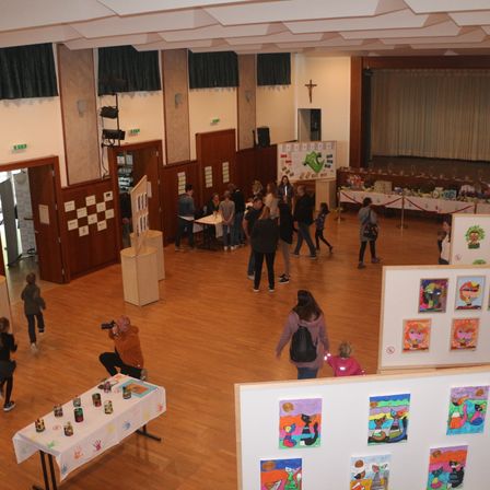 Eine Indoor-Kunstausstellung mit vielen Menschen, die herumlaufen. Bunte Gemälde an der Wand und ein Tisch mit Handabdrücken. Ein Mann macht ein Foto mit einer Kamera.
