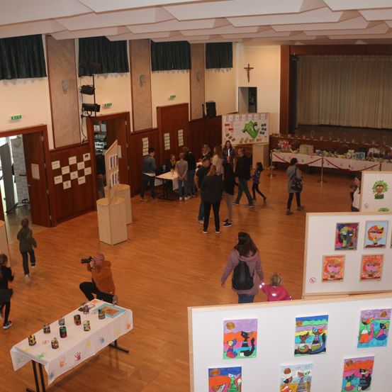Eine Indoor-Kunstausstellung mit vielen Menschen, die herumlaufen. Bunte Gemälde an der Wand und ein Tisch mit Handabdrücken. Ein Mann macht ein Foto mit einer Kamera.