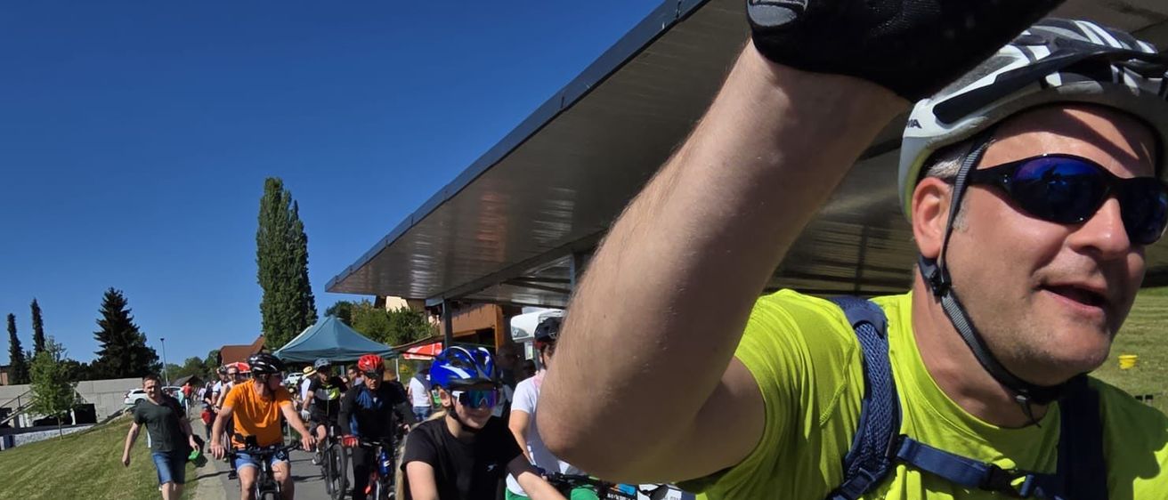 Eine Gruppe von Radfahrern fährt gemeinsam auf einem Weg unter einem Schutzdach, ein Mann winkt mit dem Arm und hält eine rote Flasche. Bäume und ein Gebäude sind im Hintergrund.