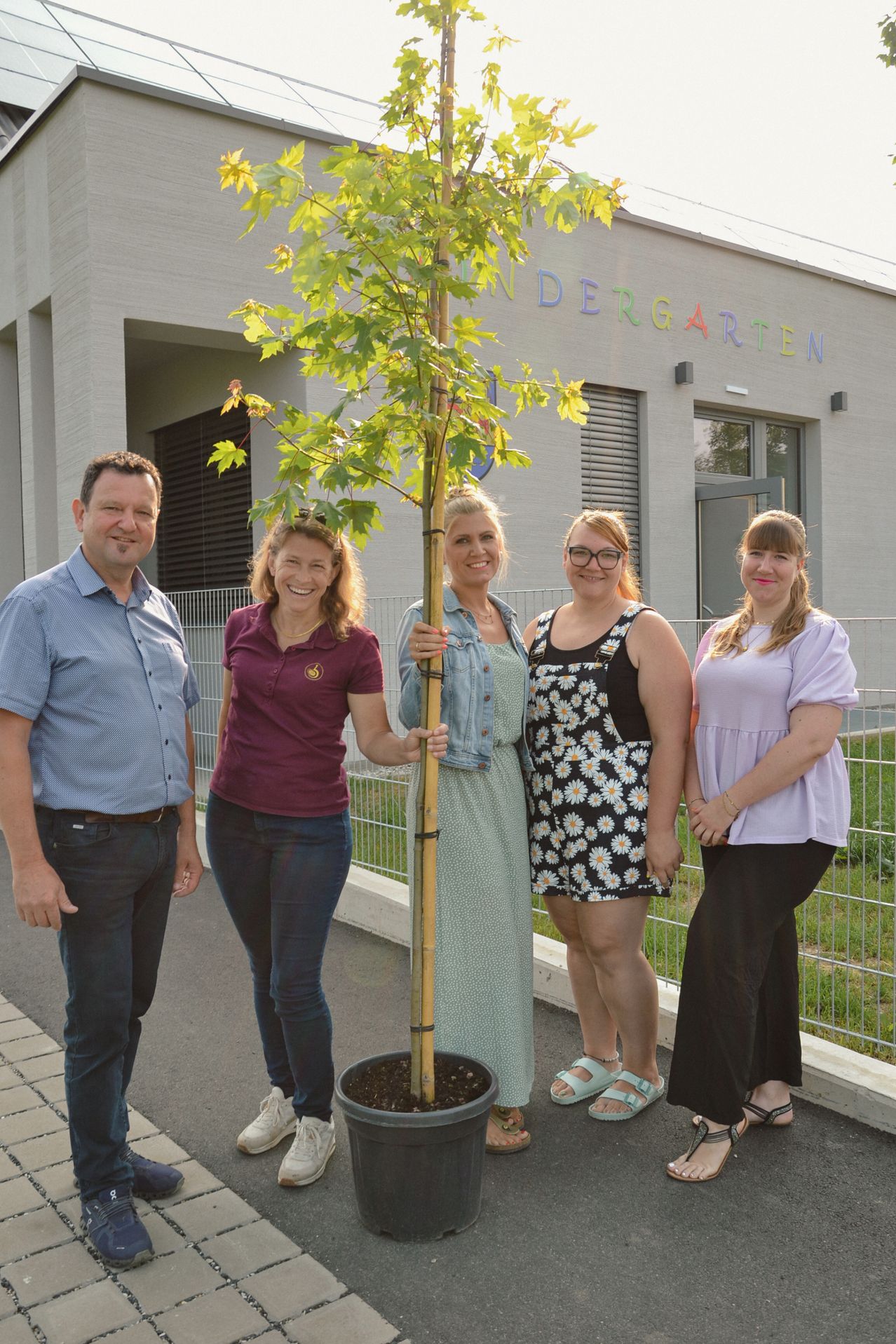 Fünf Erwachsene stehen vor einem Gebäude mit einem Baum in der Mitte. Sie alle lächeln. Das Gebäude sagt Kindergarten.