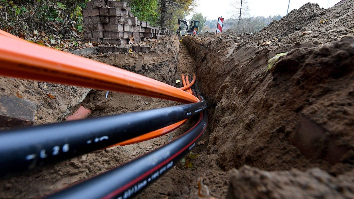 Eine Baustelle mit orangefarbenen und schwarzen Kabeln, die im Boden vergraben sind. In der Nähe steht ein Traktor, und es gibt Ziegelmauern und Bäume im Hintergrund.