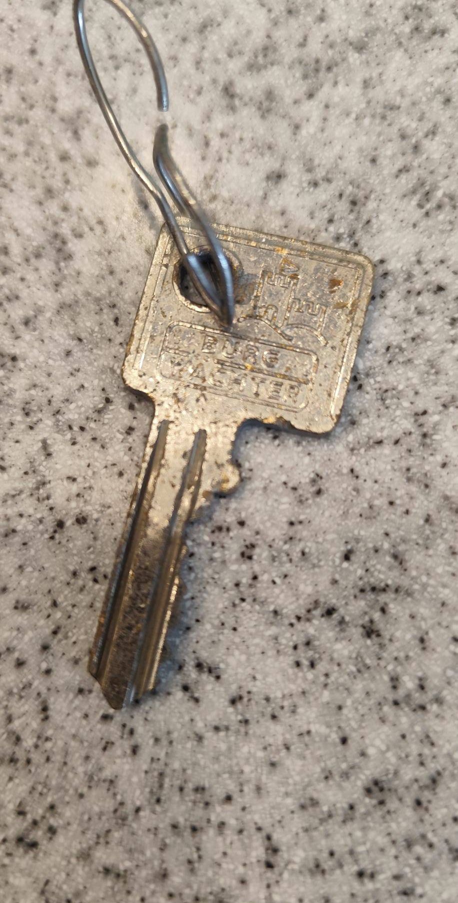 Ein alter, abgenutzter silberner Schlüssel mit der Aufschrift 'BURG MASTER', der an einem silbernen Ring befestigt ist, liegt auf einer gesprenkelten grauen Oberfläche.