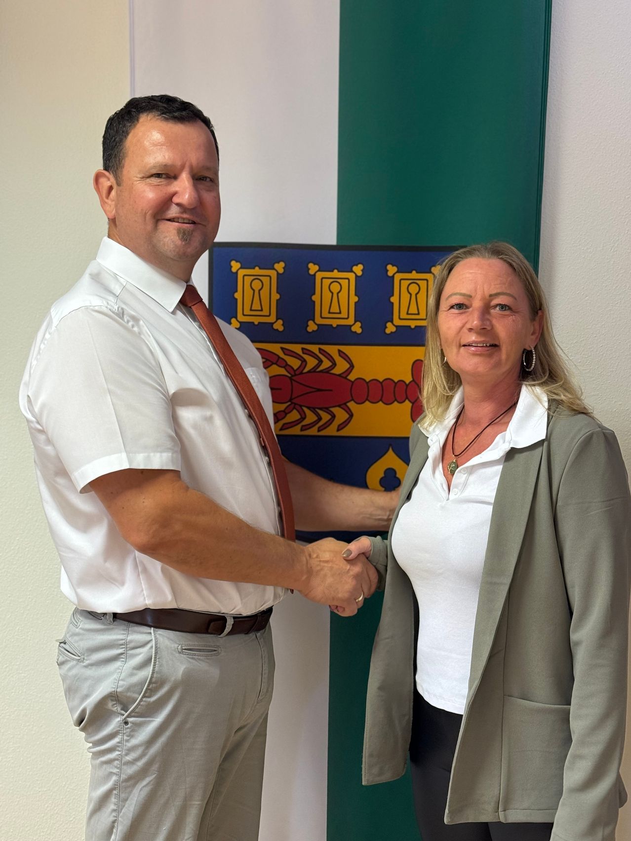 Ein Mann und eine Frau schütteln sich die Hände vor einem Wappen, beide lächeln. Der Mann trägt ein weißes Hemd, eine Krawatte und graue Hose, während die Frau ein weißes Hemd, einen Blazer und schwarze Hose trägt.