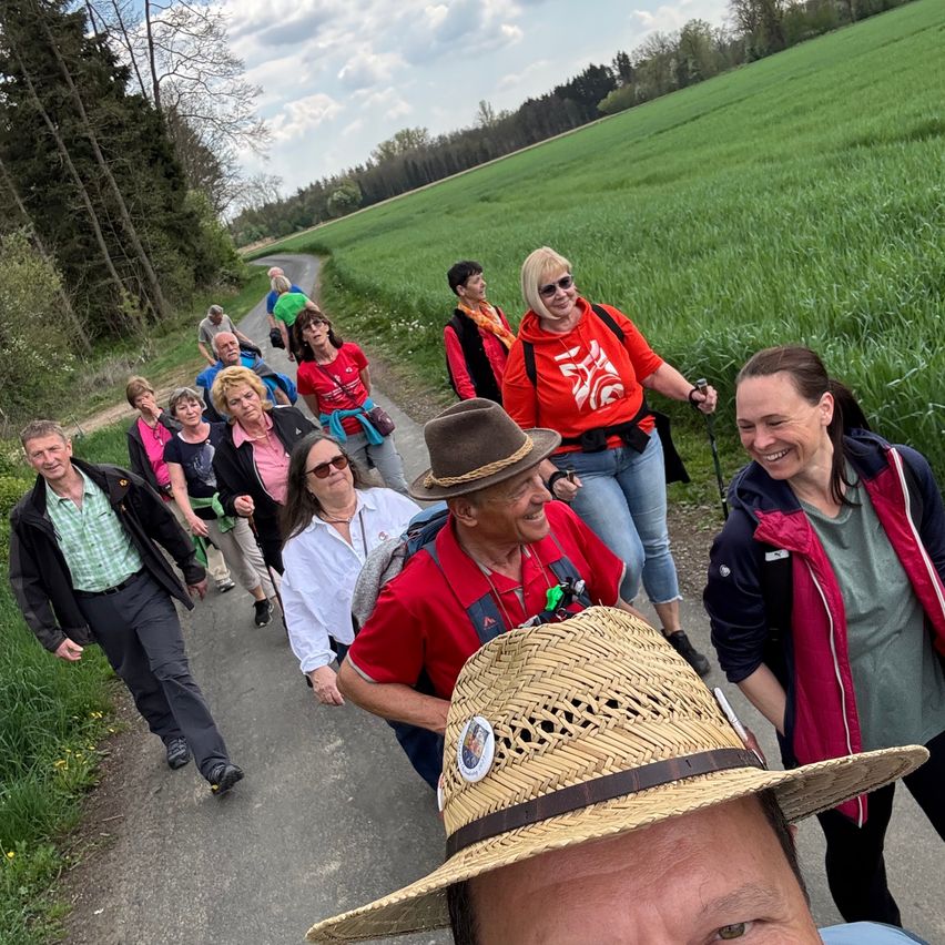 A group of people are walking in a field. Some are wearing hats and glasses. The man in the front is wearing a straw hat.