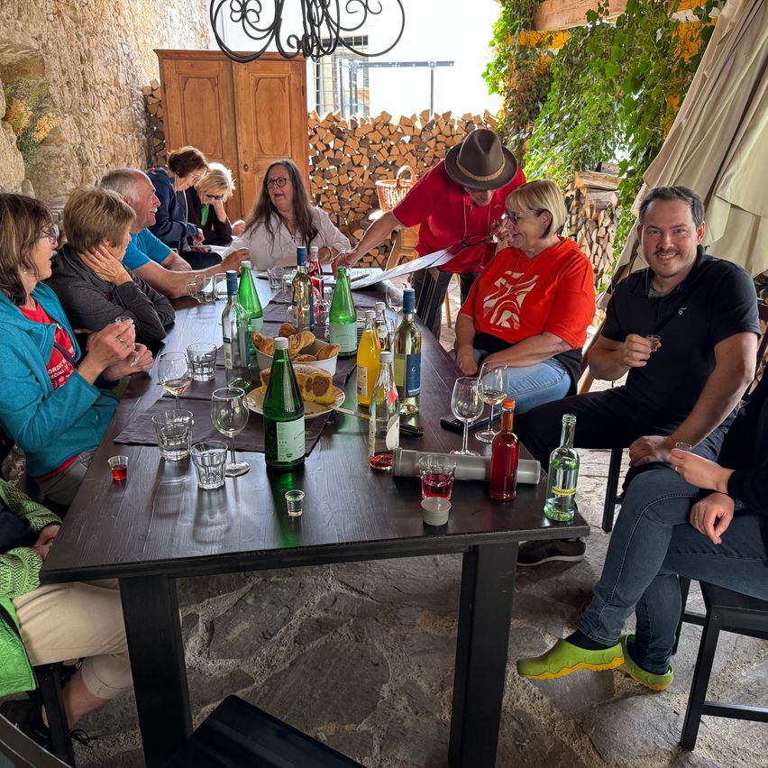 A group of people sit around a table filled with drinks and food. A man in a hat stands at the table, and another man smiles. A woman in red smiles at the camera.