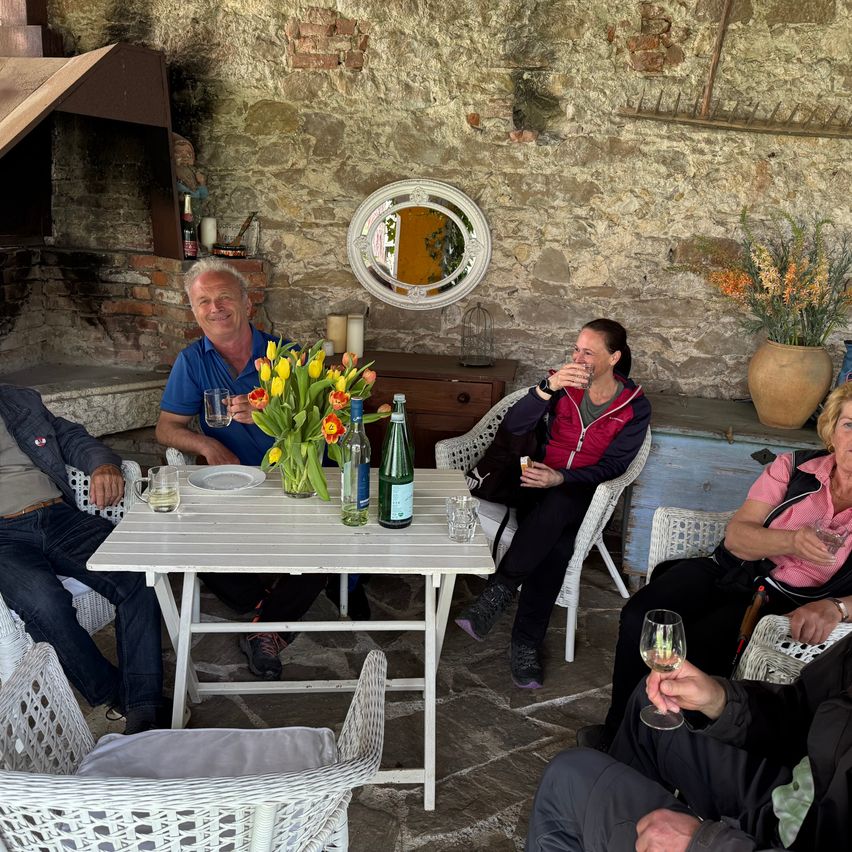 A group of people are sitting in a garden around a table with flowers and wine glasses. The man is smiling and holding a glass. The woman is drinking from a glass.