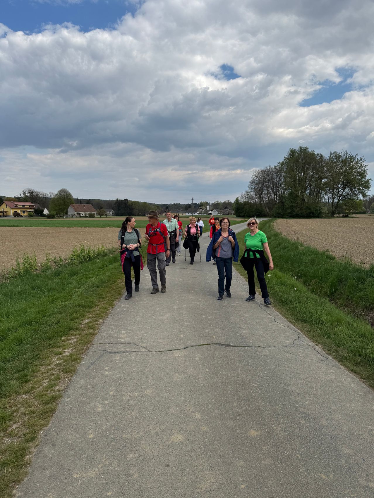 A group of people are hiking on a concrete path surrounded by grass. They are wearing casual clothes and some have backpacks. Behind them is a field with trees and houses.