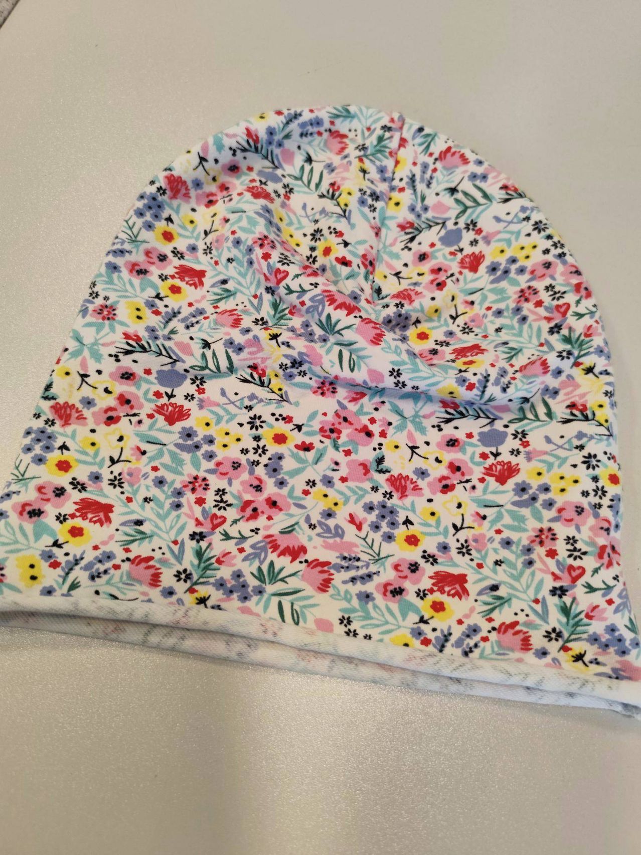 Eine bunte Mütze mit Blumendruck und weißem Band, gefaltet und auf einer weißen Oberfläche platziert.