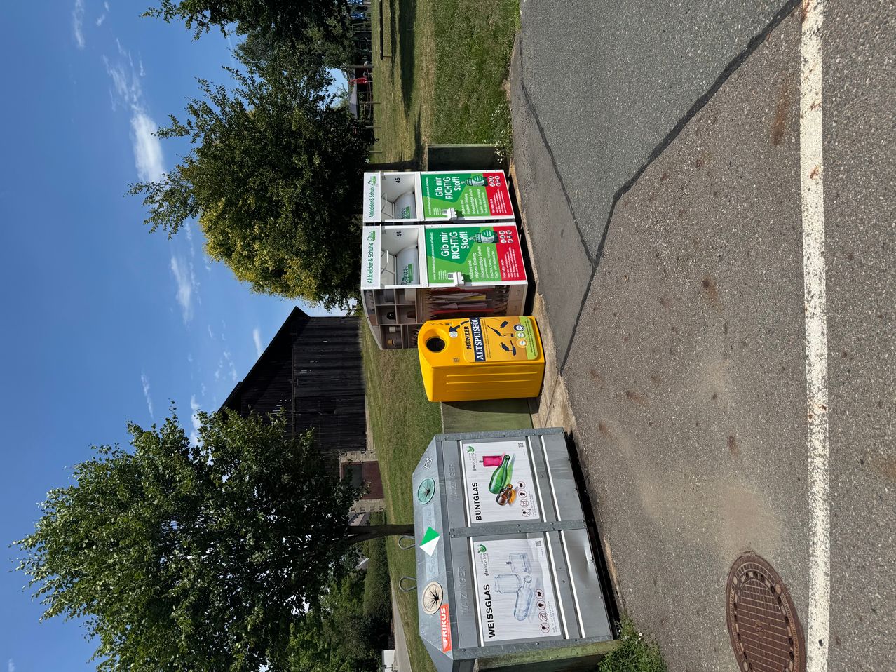 Three recycling bins are on the grass. A yellow bin for mixed waste and two grey bins for colored glass and clear glass.