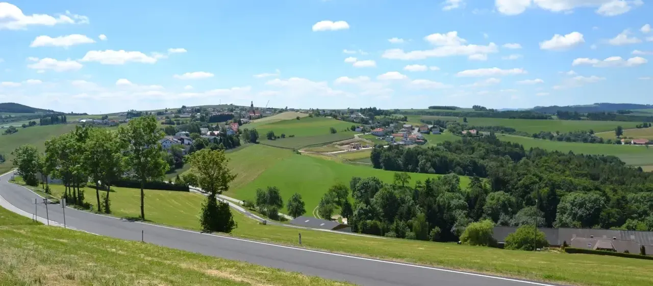 Luftaufnahme einer ländlichen Landschaft mit grünen Feldern, Bäumen und einer Straße. Ein kleines Dorf mit Häusern ist in der Ferne sichtbar. Der Himmel ist blau mit verstreuten Wolken.