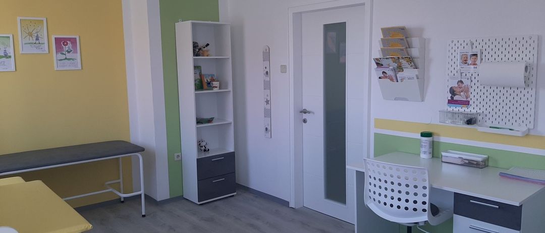 Ein kleiner Raum mit einem weißen Schreibtisch, Stuhl und Bücherregal. Der Raum hat eine weiße Tür mit Glasscheibe und einen Zeitschriftenständer an der Wand. Der Raum hat eine grün-gelbe Akzentwand.