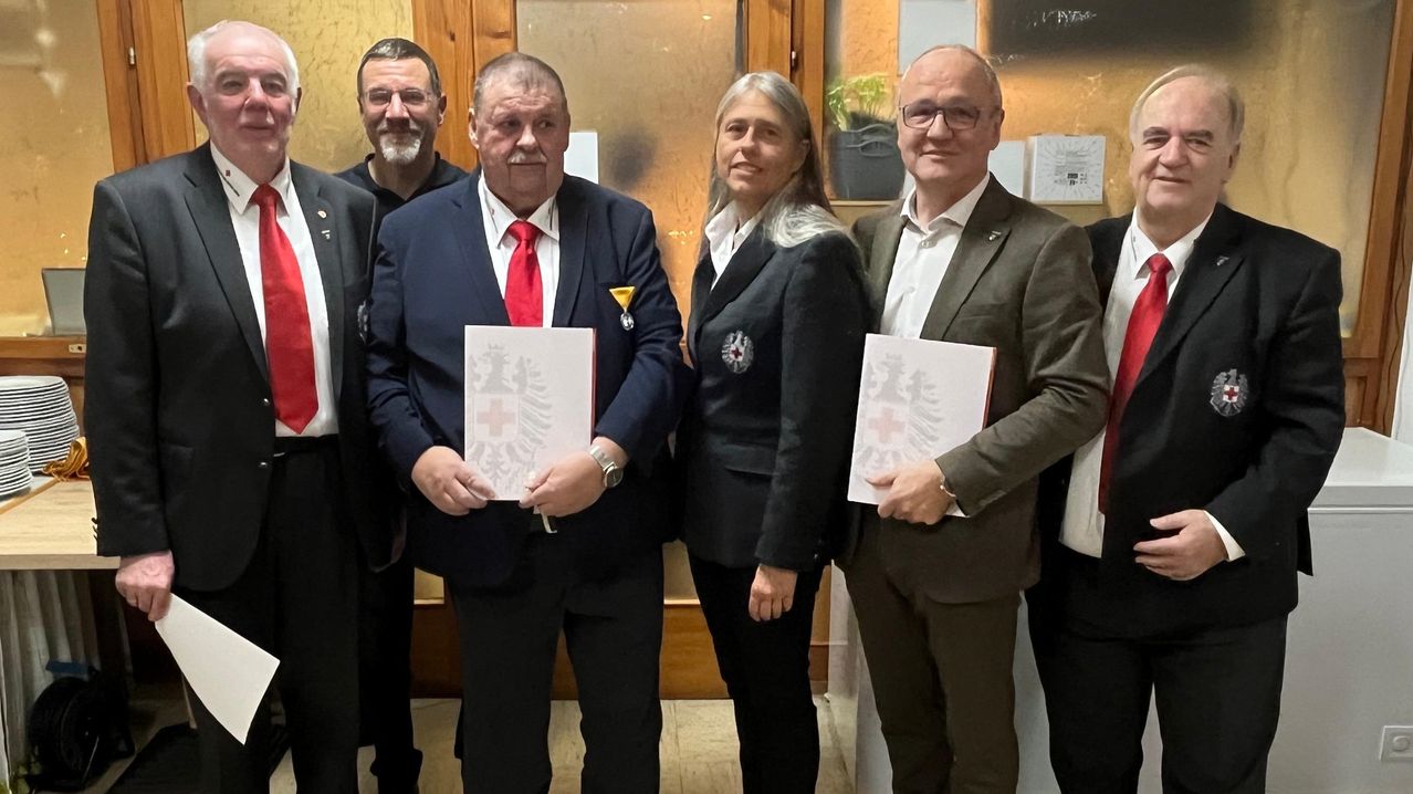 Fünf Personen, darunter ein Mann mit roter Krawatte und eine Frau mit langen Haaren, stehen nebeneinander und halten Papiere, wahrscheinlich in einer formellen Umgebung.