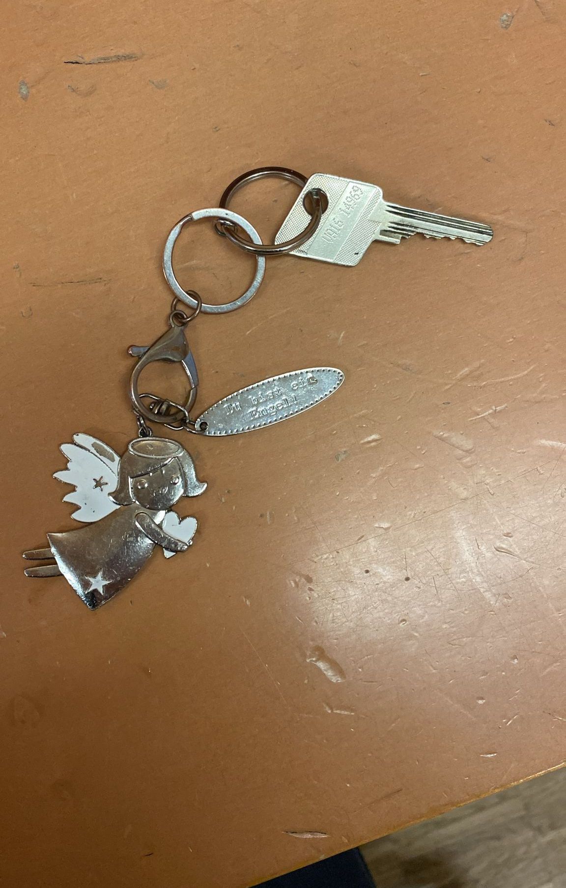 Ein silberner Schlüsselanhänger mit einem Engel, der ein Herz hält, ist an einem Schlüssel befestigt. Der Engel trägt einen Heiligenschein und Flügel. Der Schlüssel hat einen Aufkleber mit der Aufschrift 'Ich werde immer bei dir sein'.