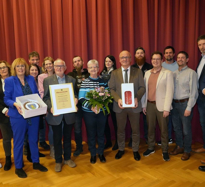 Eine Gruppe von Menschen steht auf einem Holzboden, hält Auszeichnungen und Boxen und lächelt für ein Foto vor einem roten Vorhang.