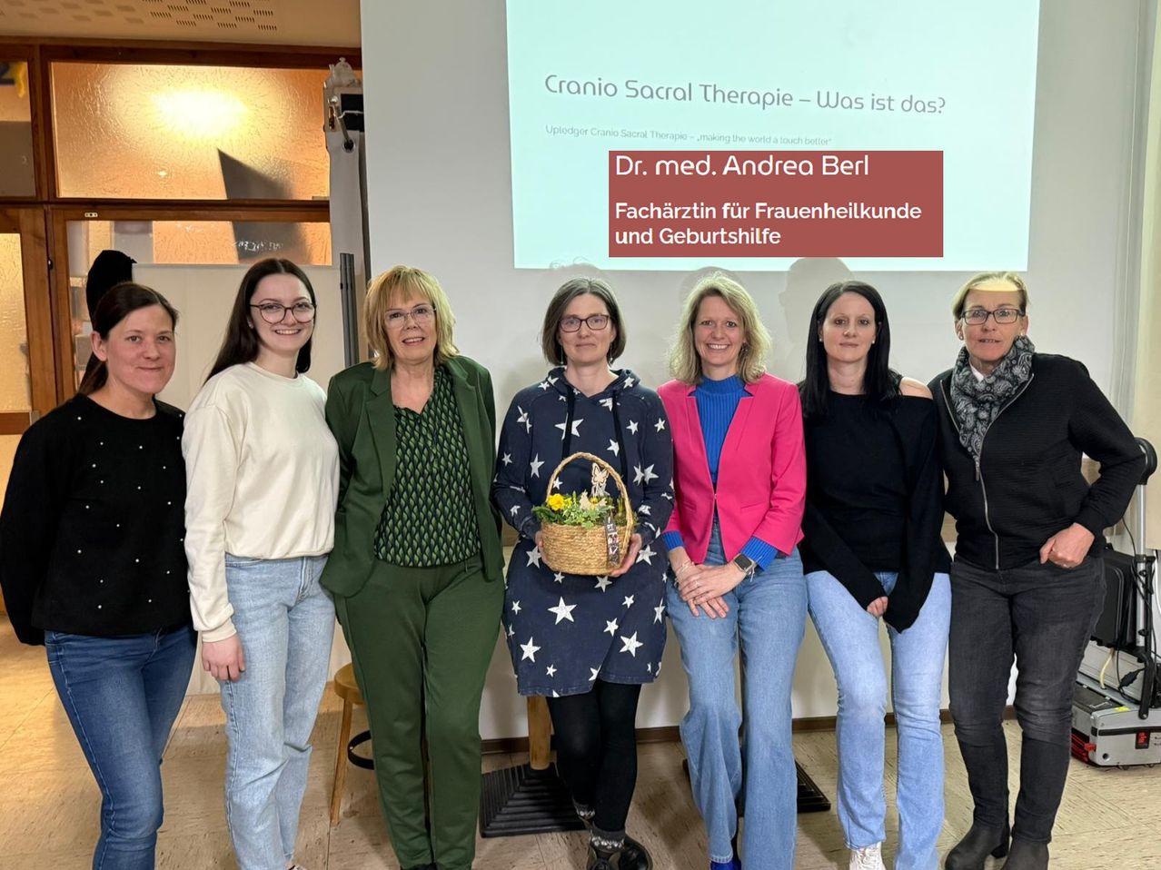 Sieben Frauen stehen vor einem Präsentationsbildschirm. Eine hält einen Korb mit Blumen. Der Präsentationstitel lautet 'Cranio Sacral Therapie - Was ist das?'.