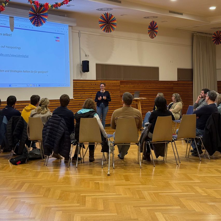 Eine Frau hält eine Präsentation in einem Klassenzimmer vor einer Gruppe von Menschen, die in Stühlen sitzen. Eine Projektionsleinwand zeigt eine Folie mit einem deutschen Text.