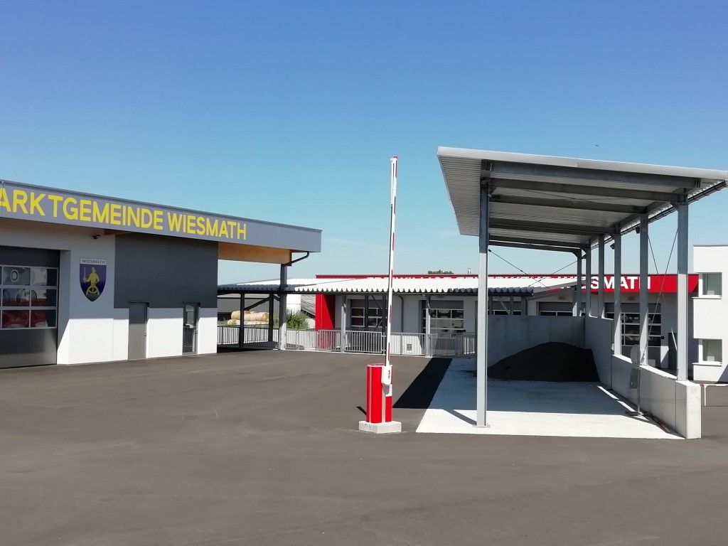 Ein Außenbereich zeigt ein Gebäude mit der Aufschrift 'Meinde Wiesmath' und einen überdachten Autowaschplatz mit einer Schranke. Der Himmel ist klar und blau.