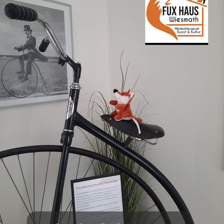 Ein altes Fahrrad mit einem Fuchs-Plüschtier auf dem Sattel, ausgestellt in einem Museum mit einem Schwarz-Weiß-Foto eines Mannes, der ein Fahrrad fährt, im Hintergrund.