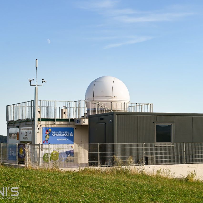 Ein modernes Gebäude mit einer großen Kuppel auf der Spitze und einem blauen Schild mit der Aufschrift 'Wiener Neustadter Sparkasse'. Das Gebäude ist von einem Zaun umgeben und befindet sich in einem Grasbereich.