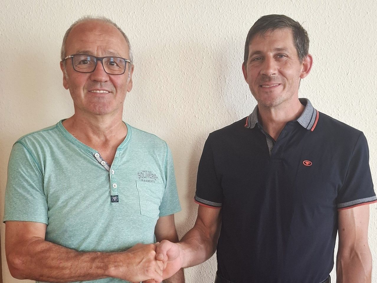Zwei Männer, einer mit Brille und hellgrünem Shirt, der andere in einem dunkelblauen Polohemd mit Logo, stehen eng beieinander und lächeln für ein Foto.