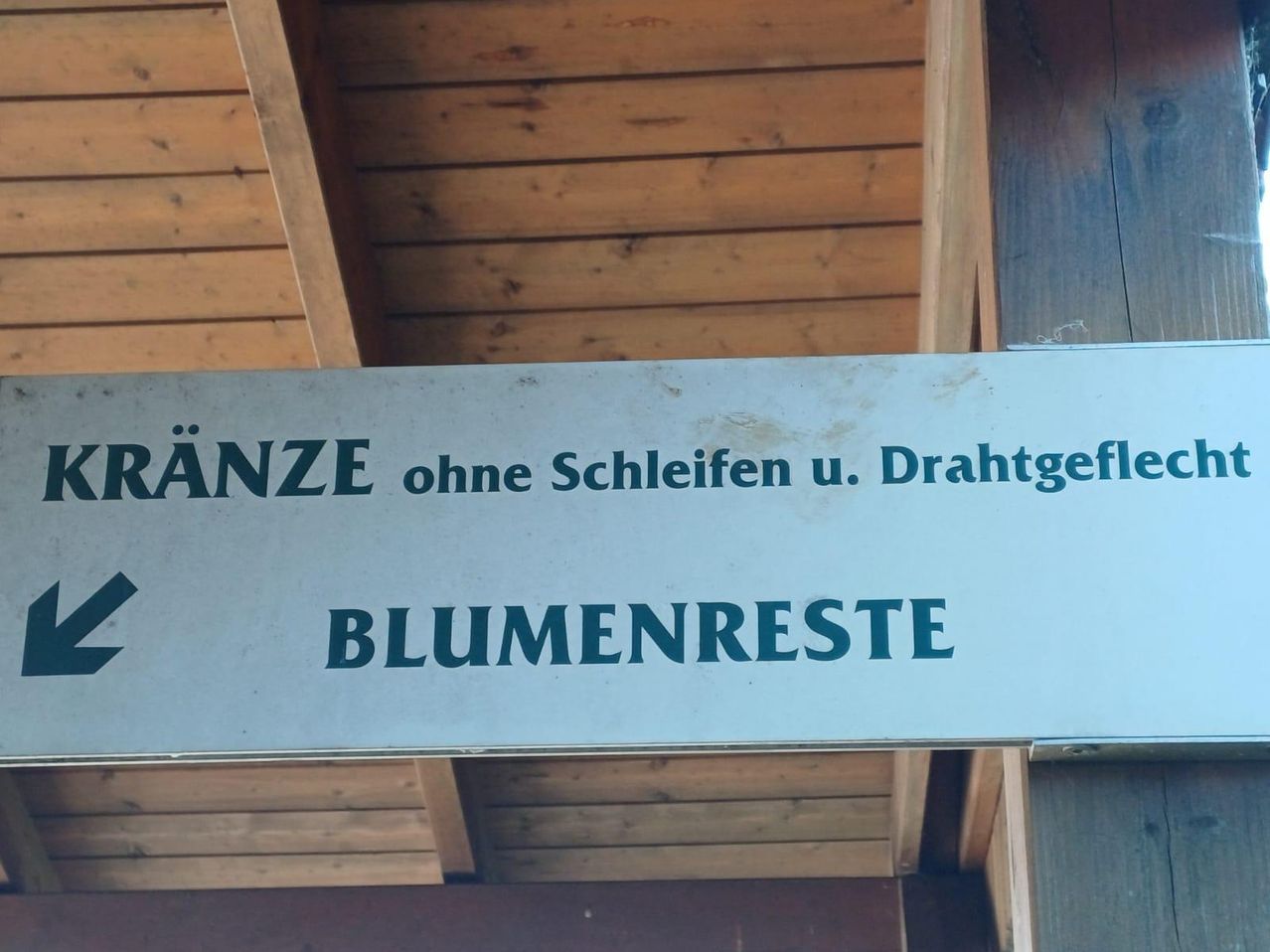 Ein Schild mit grüner Schrift auf weißem Hintergrund hängt von einer Holzdecke. Der Text lautet 'ANZE ohne Schleifen u. Drahtgef BLUMENRESTE'.