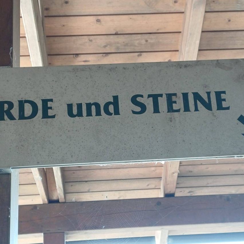 Ein Betonschild mit den Worten 'ERDE und STEINE' in Schwarz auf einer Holzdecke. Das Schild ist an der Decke befestigt.