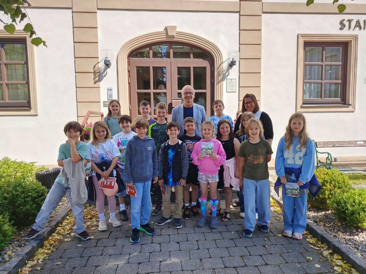 Eine Gruppe von Kindern und Erwachsenen steht vor einem Gebäude mit einer großen Bogentür. Die Kinder halten Bücher und scheinen für ein Foto zu posieren.