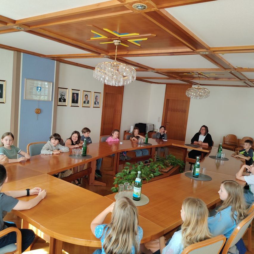 Ein Klassenzimmer mit Schülern und Lehrern, die um einen Tisch herumsitzen, mit zwei Kronleuchtern an der Decke. Es gibt Bilderrahmen an der Wand und eine Pflanze in der Mitte.