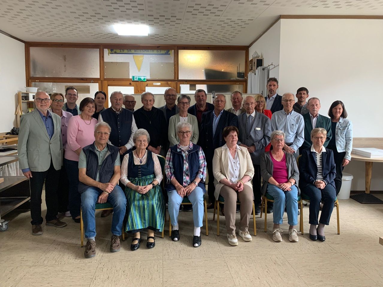 Eine Gruppe älterer Menschen posiert für ein Foto in einem Raum mit Glasfenstern und Holzrahmen. Einige von ihnen sitzen auf Stühlen, während andere hinter ihnen stehen.