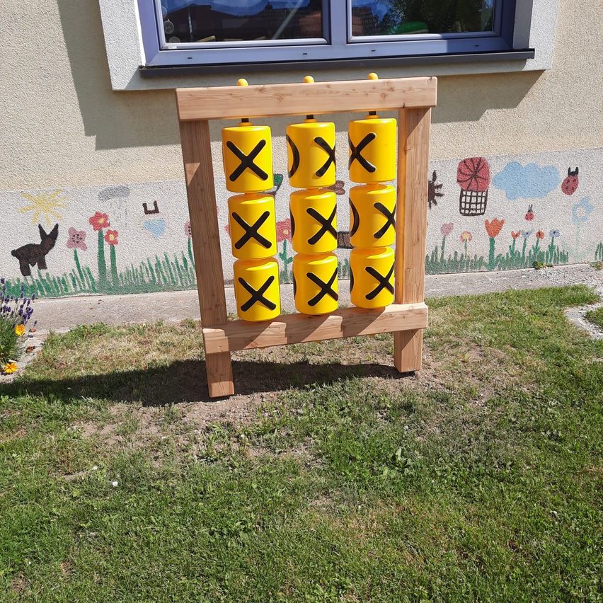 Ein hölzernes Spielsetup mit gelben Würfeln mit schwarzen X's steht vor einer Wand mit einem gemalten Wandbild.