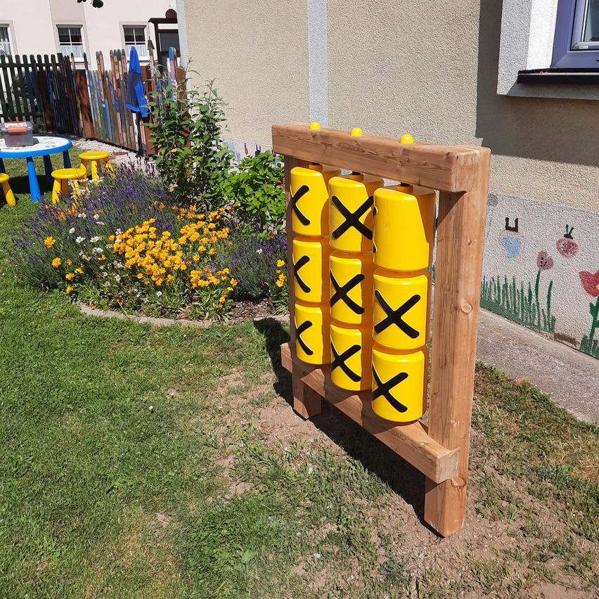 Eine hölzerne Struktur mit gelben Fässern und schwarzen X-Markierungen im Freien, in der Nähe eines Gartens mit Blumen und einem Haus.