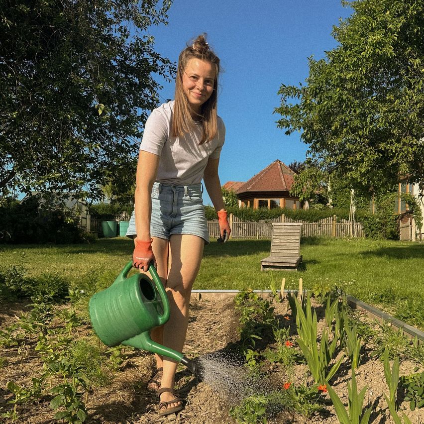 Eine Frau gießt Pflanzen in einem Garten mit einem grünen Gießkannen, trägt Handschuhe und Sandalen. Hinter ihr befindet sich ein Haus mit einem Holzzaun und einer Bank.