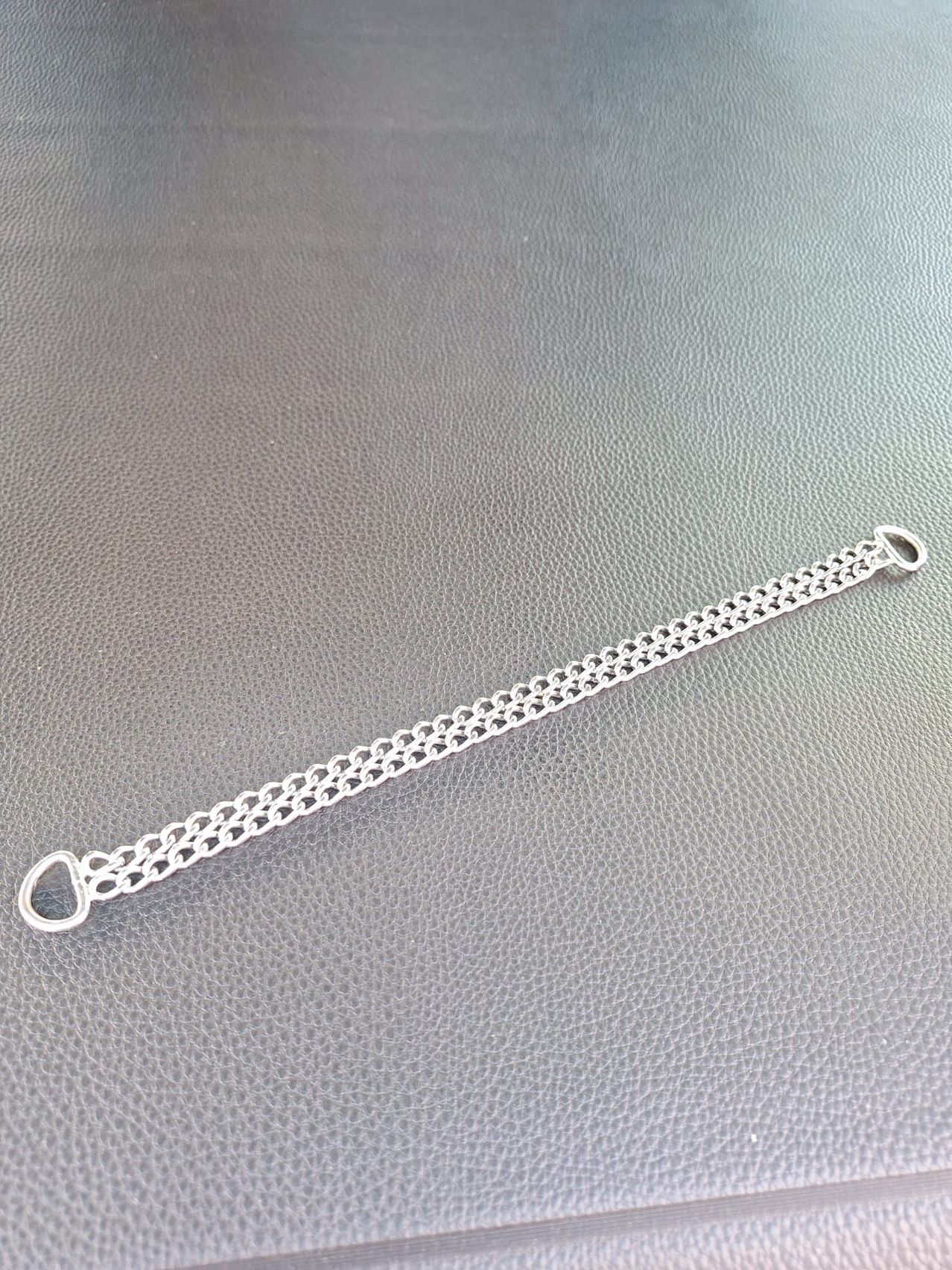 Ein silberner Kettenarmreif mit herzförmiger Schließe liegt auf einer grauen Lederoberfläche. Die Kette hat ein zartes Design.