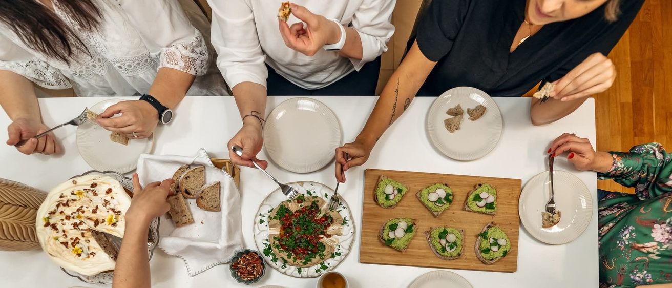 Eine Gruppe von Menschen sitzt um einen Tisch und isst. Der Tisch ist mit Tellern, Schalen und einem Holzschneidebrett mit Essen darauf bedeckt.