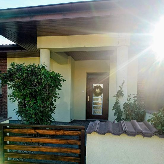 Ein modernes Haus mit einem großen Fenster und einer Tür mit einem Kranz, umgeben von Grünpflanzen und in Sonnenlicht gebadet.