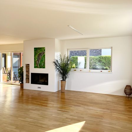 Ein leerer Raum mit weißen Wänden, Holzböden und einem Kamin. Ein großes Fenster an der Wand mit Blick nach draußen.