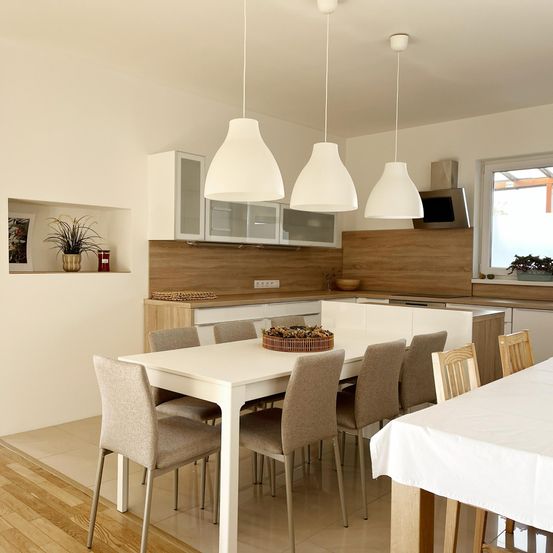 Eine moderne Küche mit Essbereich. Ein weißer Esstisch mit Stühlen ist aufgebaut, umgeben von Schränken und Holzregalen. Ein Korb mit Essen steht auf dem Tisch. Drei weiße Lampen hängen von der Decke.