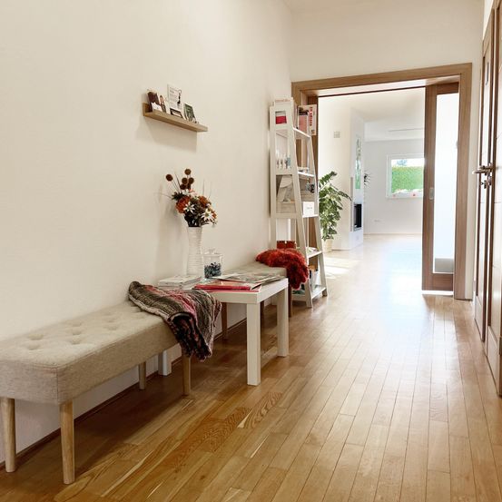 Ein Flur mit Holzboden, mit einer Bank, einem Tisch mit Blumen und Büchern und einem Regal mit Büchern. Es gibt eine Glastür zu einem anderen Raum mit einem Fenster.