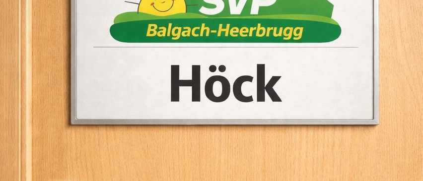 Ein Schild, das an einer Holztür befestigt ist, zeigt 'SVP Balgach-Heerbrugg Hock'. Das Logo besteht aus einer Sonne mit einer Schweizer Flagge oben und einem grünen Hut.