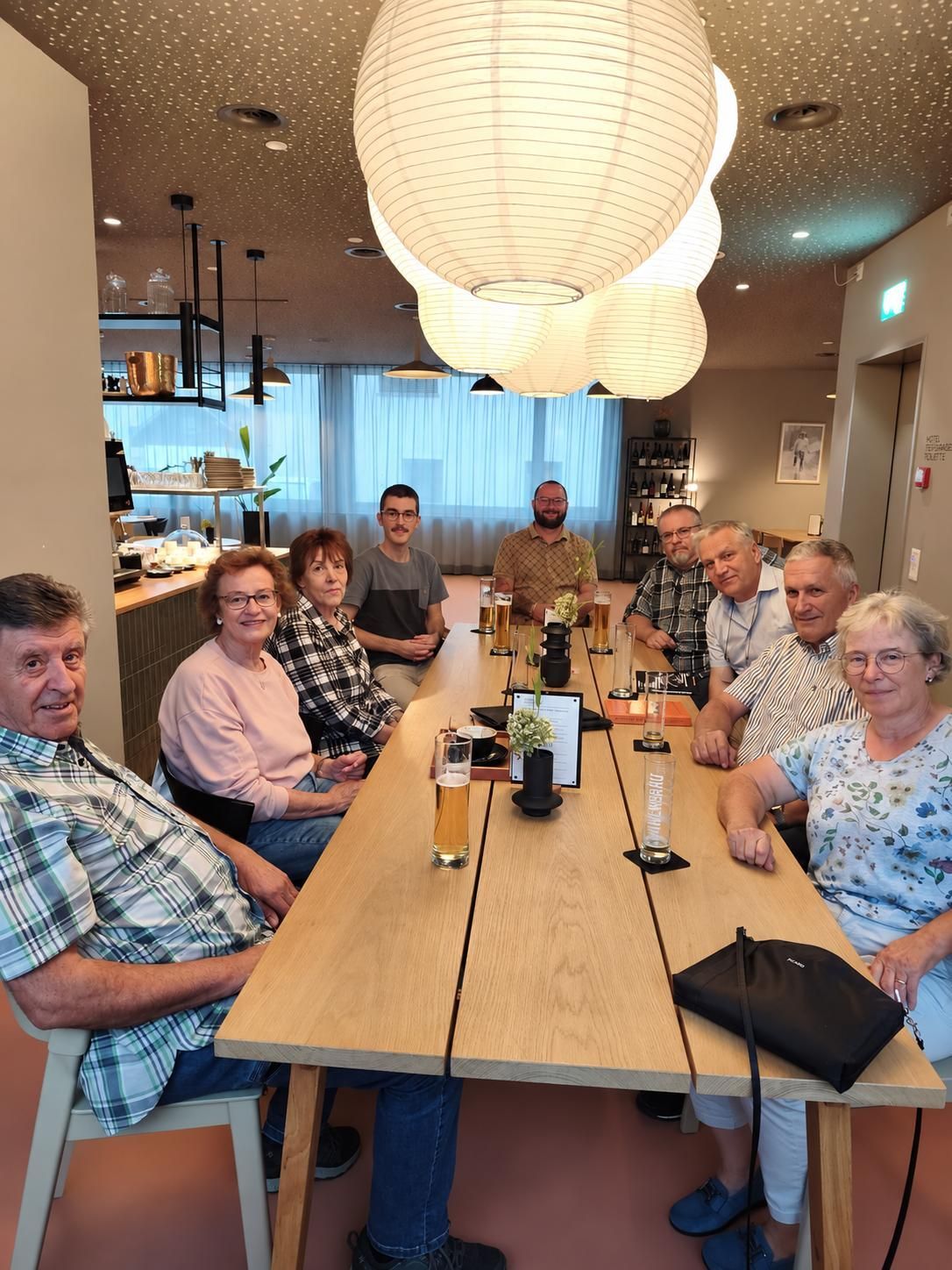 Eine Gruppe von Menschen sitzt um einen langen Tisch in einem Restaurant, lächelt und posiert für ein Foto. Sie haben Gläser mit Bier und eine Blumenarrangement auf dem Tisch. Der Raum hat weiße Vorhänge, Regale mit Flaschen und Papierlaternen, die von der Decke hängen.